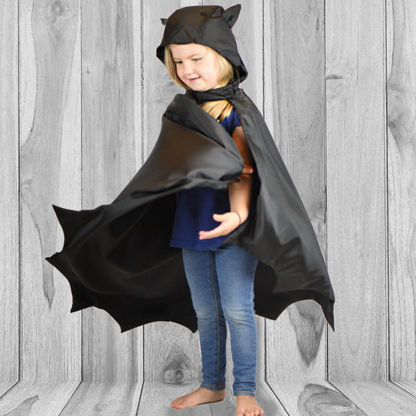 Small child with long blonde hair is twirling a black bat-shaped cape.  It has small stuffed ears on the hood of the cape.. Elastics on wrists. Child is standing on a faux-wood background, wearing jeans and t shirt.